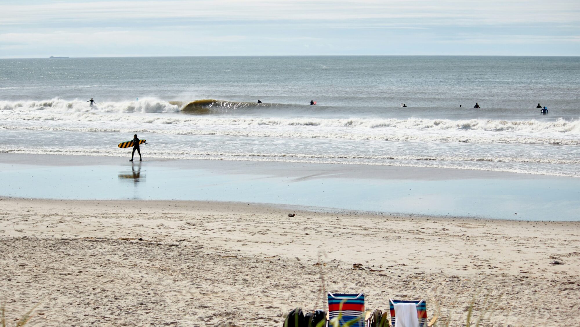 A small barrelling wave seen from the shore as a surfer with a malibu exits the water; two empty beach chairs rest on the sand at Rockaway Beach surf, Queens, NY