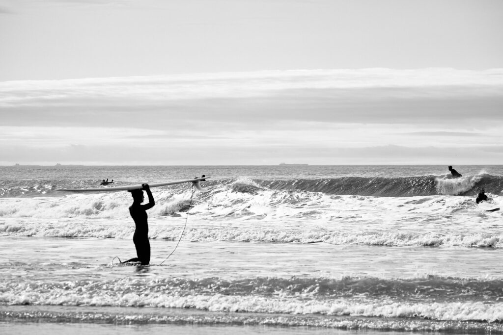 Surfer walks into the ocean at Rockaway Beach surf, Queens, NY, carrying a Malibu board overhead, with a wave breaking in the background in black and white