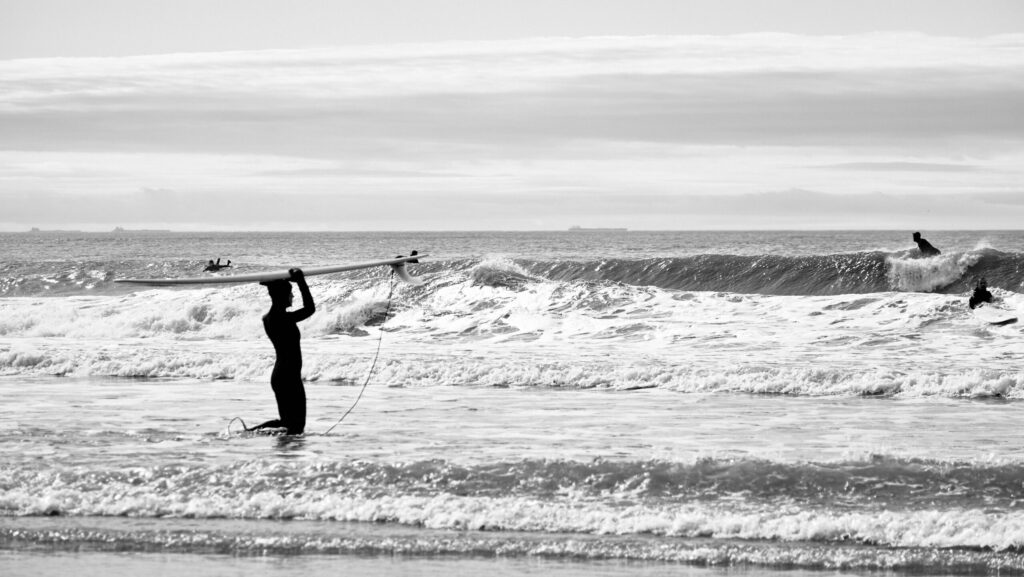 Surfer walks into the ocean at Rockaway Beach surf, Queens, NY, carrying a Malibu board overhead, with a wave breaking in the background in black and white