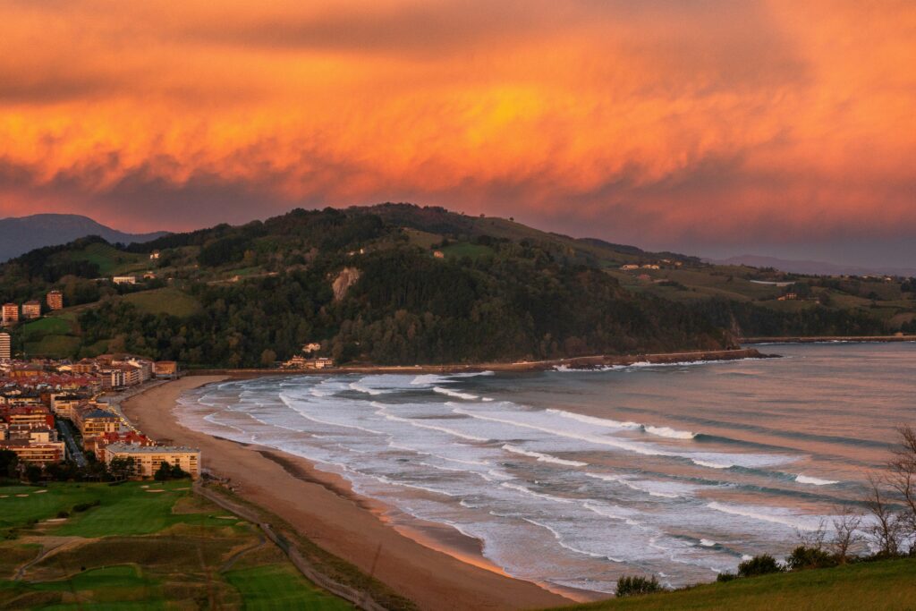 Sunset view of Zarautz Surf Bay, Spain Surf, with rolling surf lines, glowing orange clouds, and a rolling hill in the background