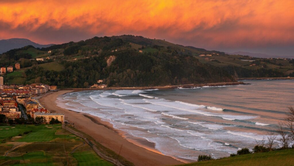 Spain Surf Guide 2025 | Best Surf Towns With Easy Access Sunset view of Zarautz Surf Bay, Spain Surf, with rolling surf lines, glowing orange clouds, and a rolling hill in the background