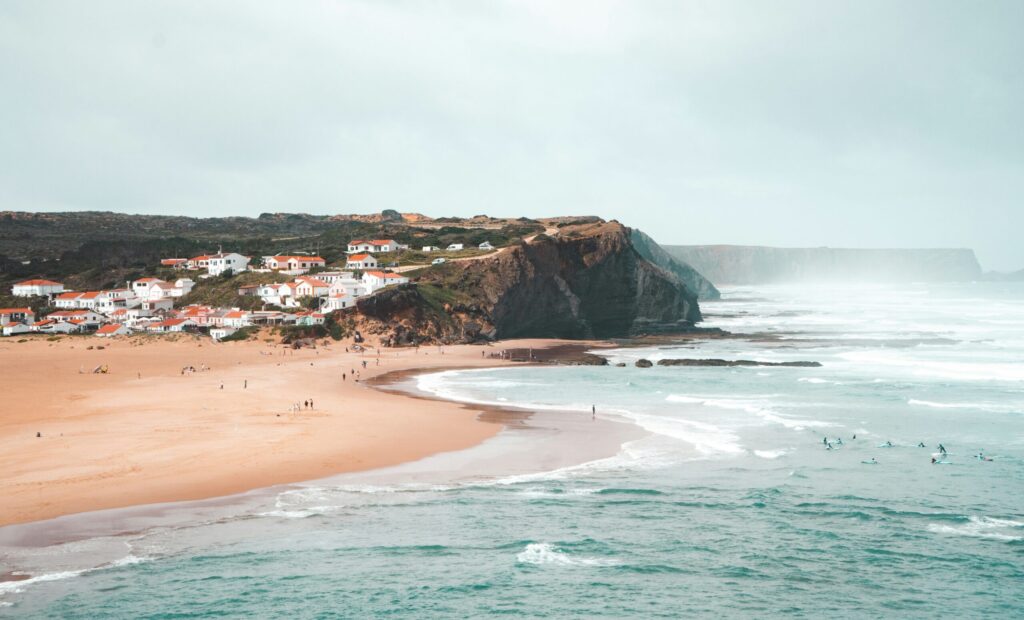 A scenic view of the cliff face and the town of Arrifana surf in the background, featuring the ocean where people are learning to surf