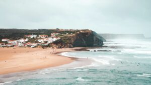 A scenic view of the cliff face and the town of Arrifana surf in the background, featuring the ocean where people are learning to surf