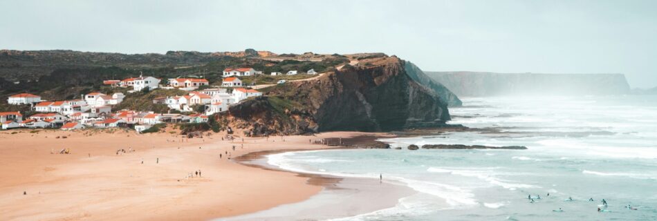 A scenic view of the cliff face and the town of Arrifana surf in the background, featuring the ocean where people are learning to surf