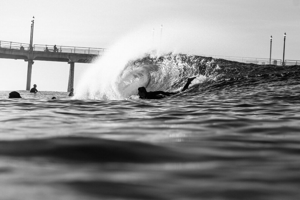 A black-and-white water shot captures a surfer paddling into a barreling wave, with the iconic Huntington Beach Surf Pier visible in the background