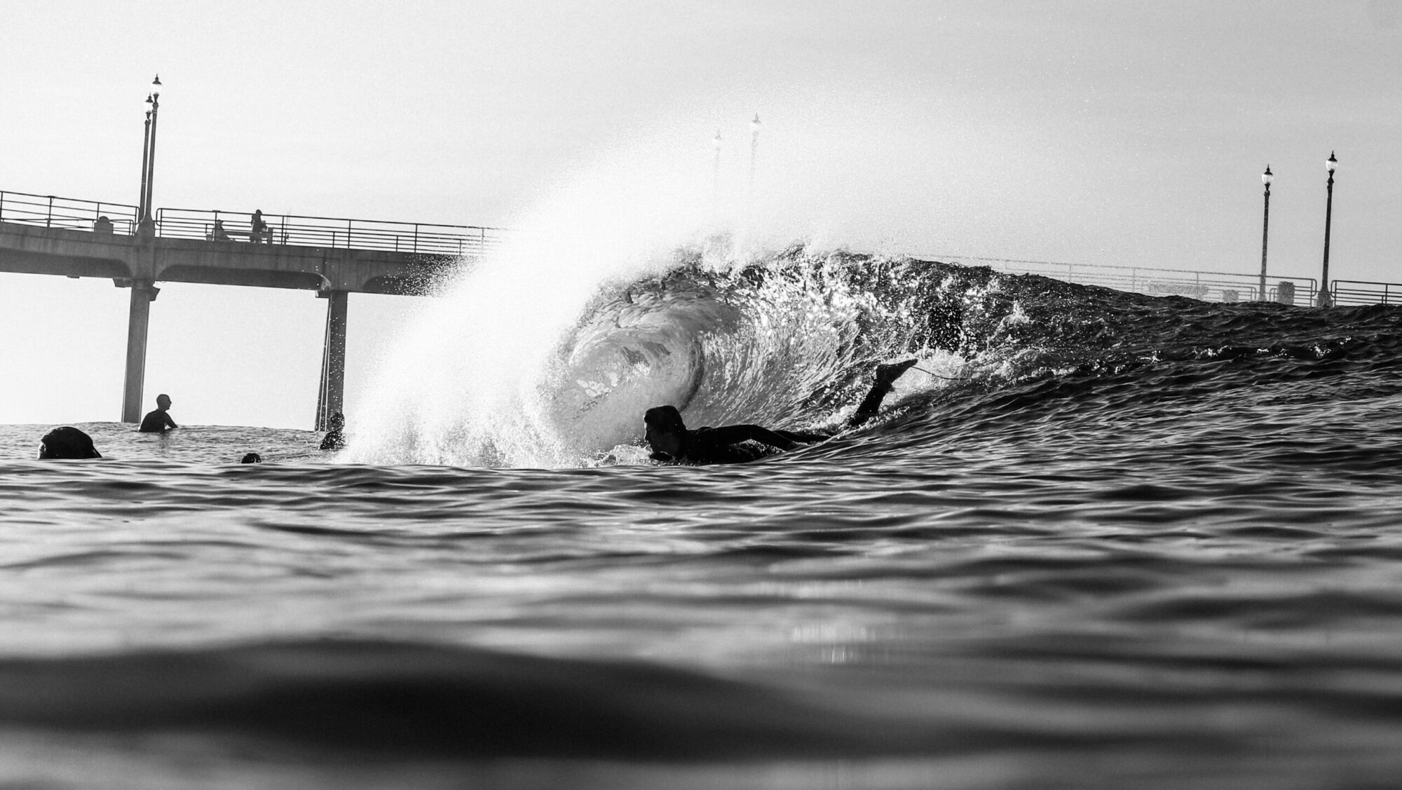 A black-and-white water shot captures a surfer paddling into a barreling wave, with the iconic Huntington Beach Surf Pier visible in the background