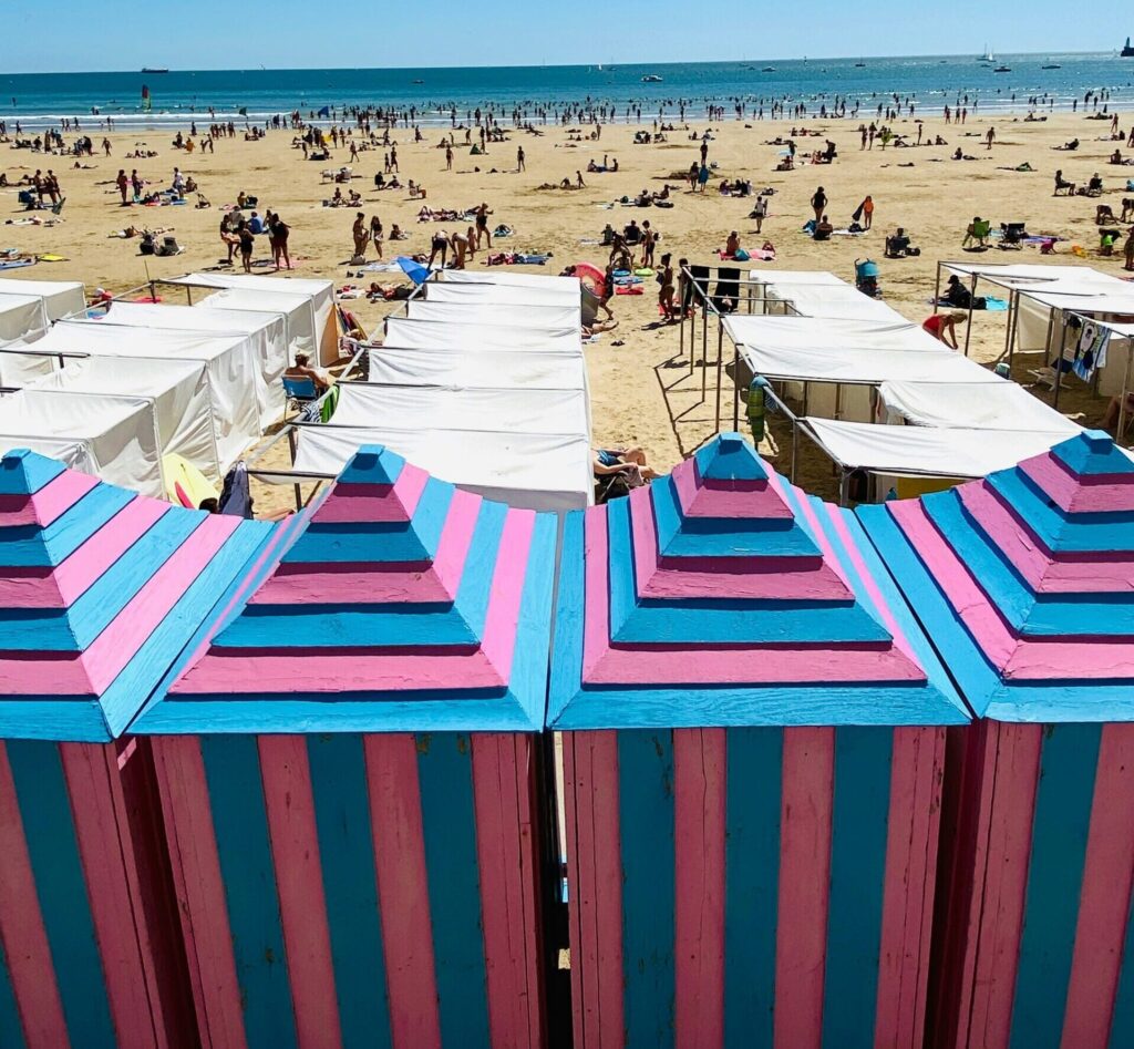 Les Sables-d'Olonne beach in France showcases bright blue and pink beach sheds, with people lounging on the sand under the sun