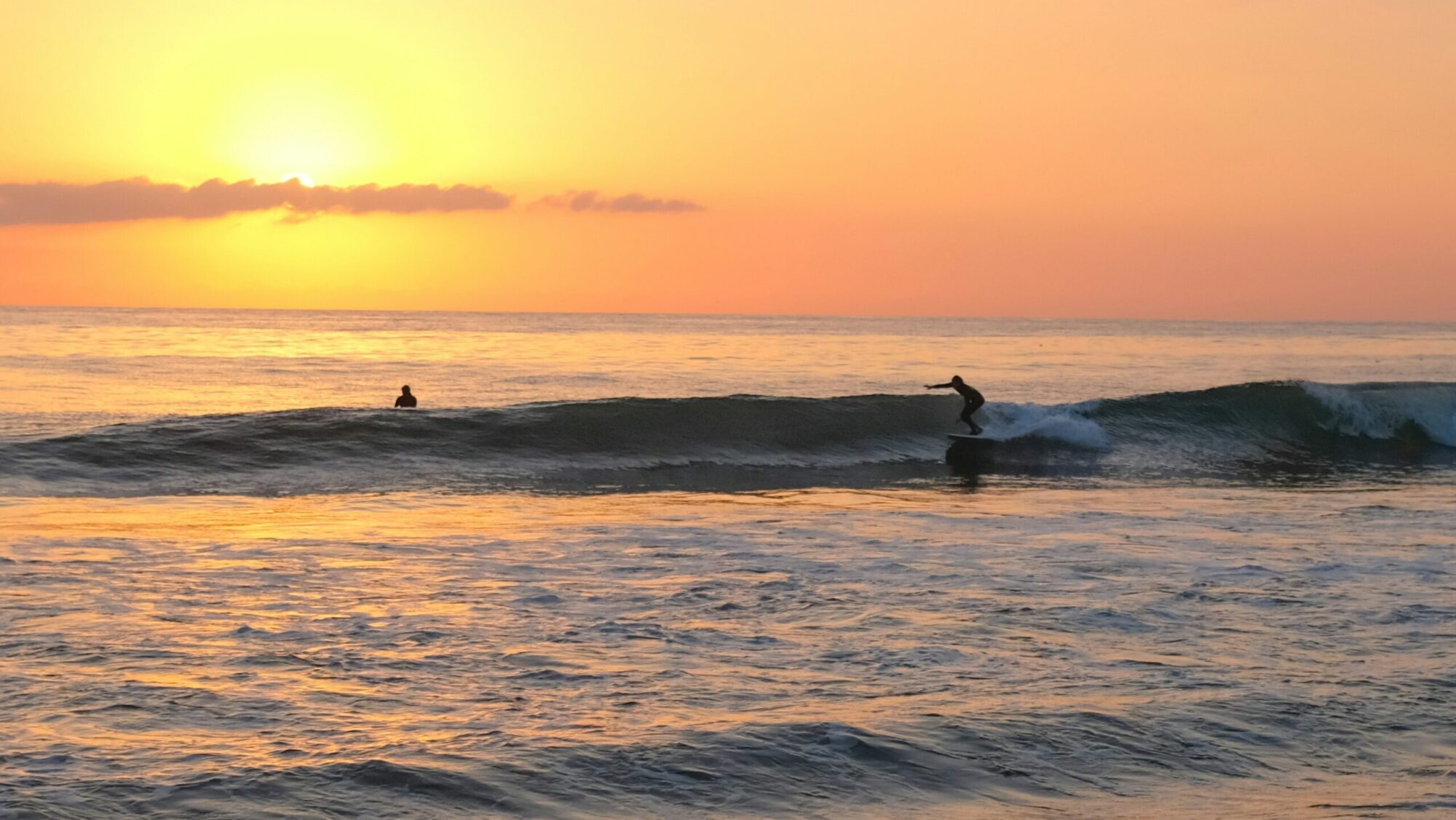 Surfer racing along a mellow right hand wave at Malibu Surf, CA, with a vibrant sunset and clear yellow-orange skies in the background