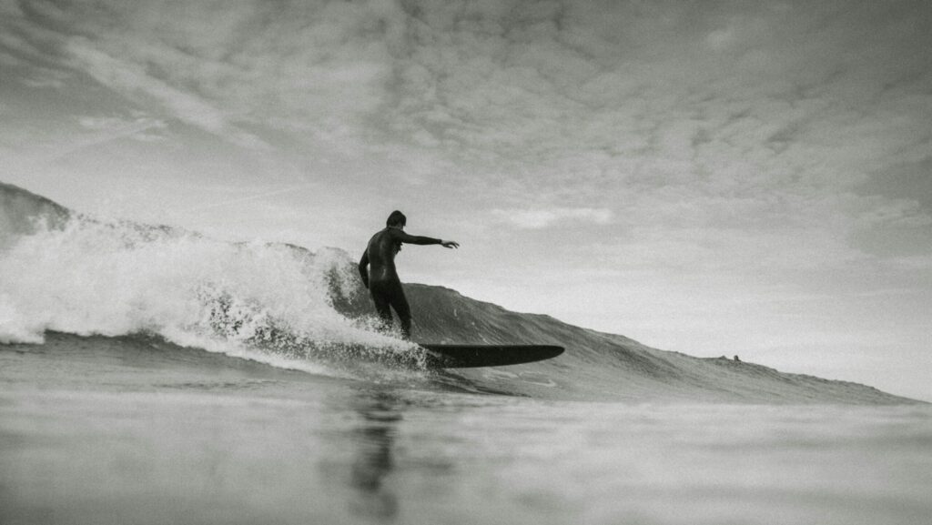 A man surfing a longboard on a gentle wave at Higgins Beach surf, Maine, under a clear sky with calm ocean waters in the background.