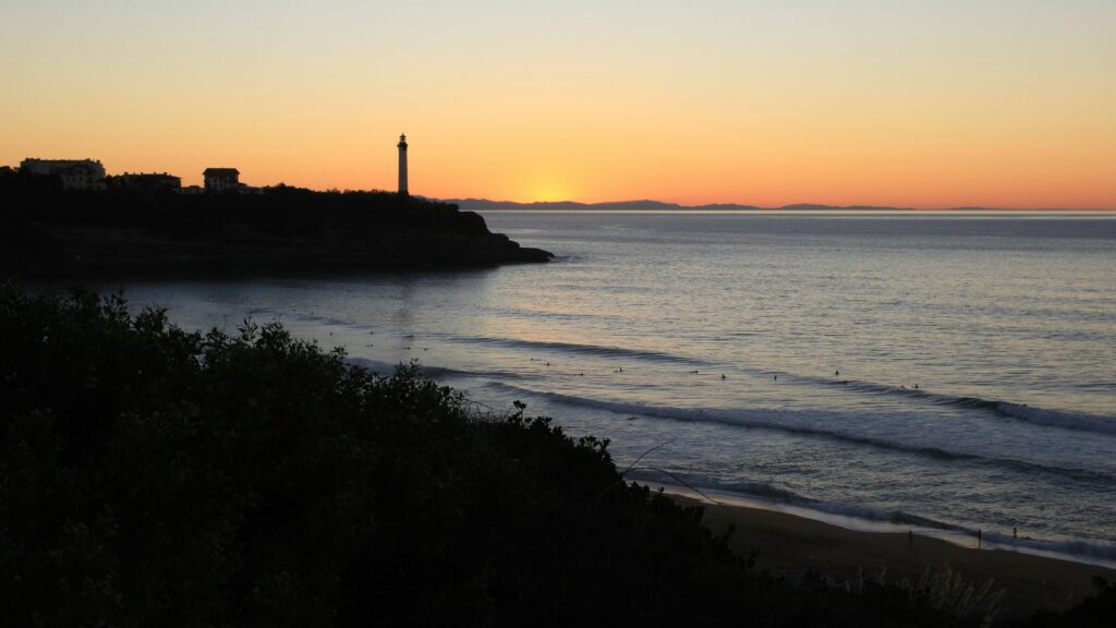 Ocean filled with surfers, with a lighthouse in the background during sunset, featuring vibrant orange and yellow skies