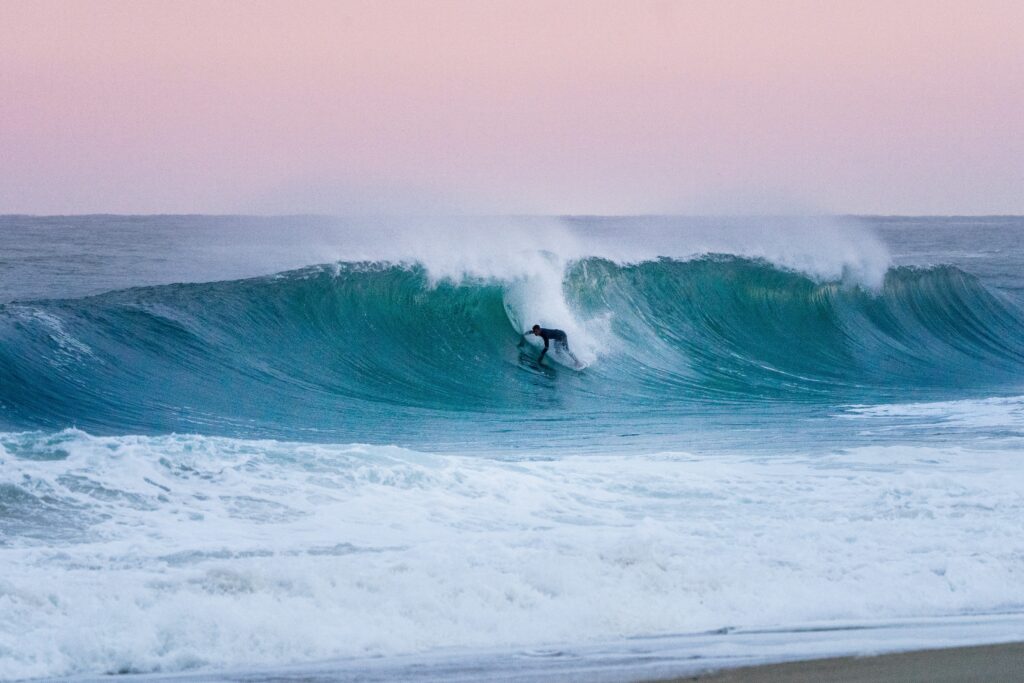 A surfer has just dropped in on a wave at Plage de la Gravière in Hossegor surf, trying to enter the barrel against a backdrop of pink and red skies.