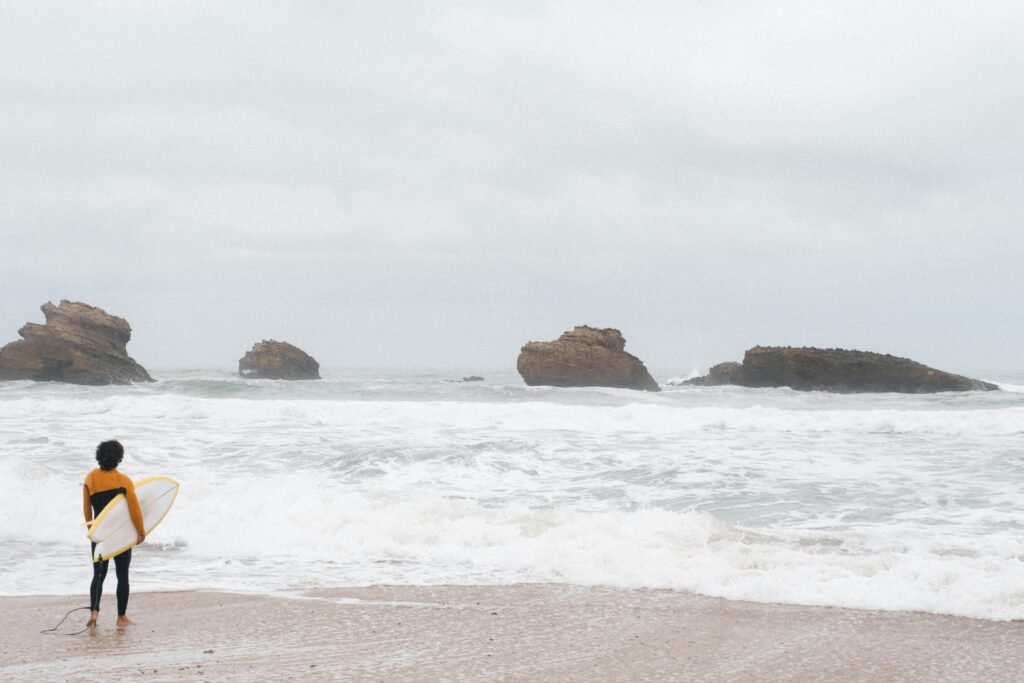 A surfer gazing out into Biarritz surf, trying to read the waves, with rocky formations in the background and grey skies