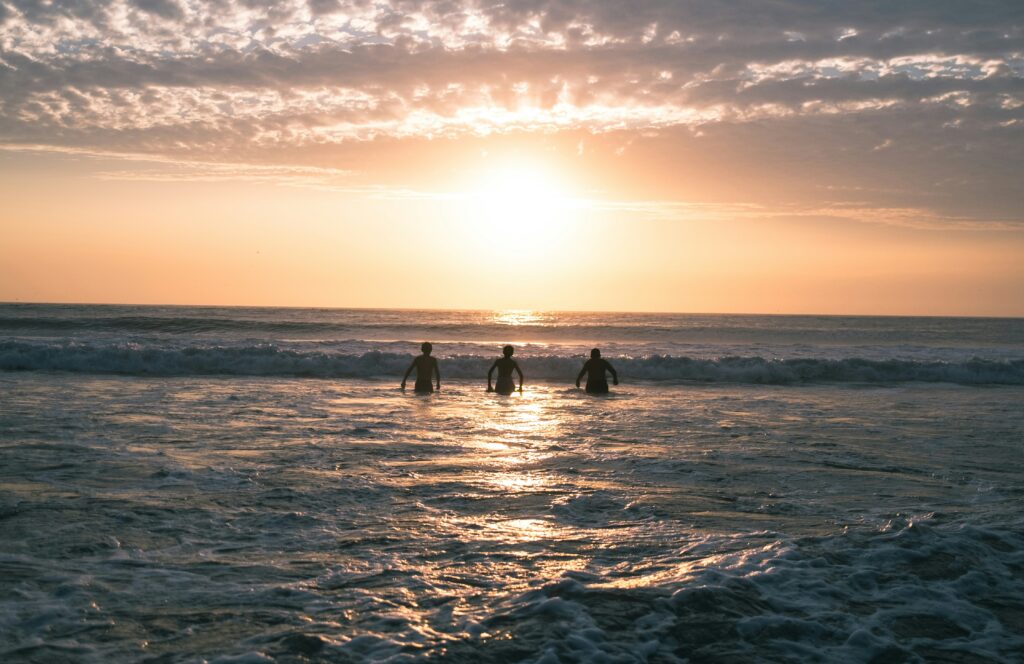 Three people entering the ocean in Miraflores, Lima, Peru surf, against a stunning sunset with vibrant yellow skies illuminating the ocean.