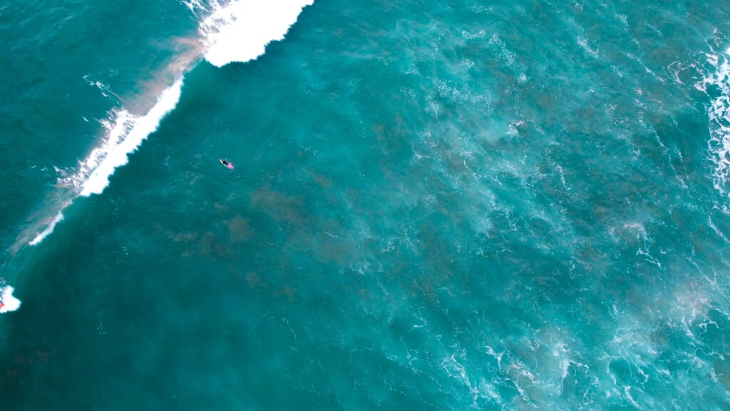 A bird's-eye view of a single surfer in blue water in Montanita surf, captured from a distance, with a wave breaking in front of them