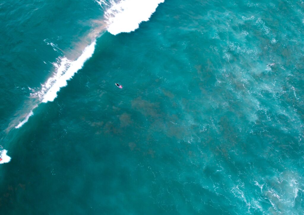 A bird's-eye view of a single surfer in blue water in Montanita surf, captured from a distance, with a wave breaking in front of them