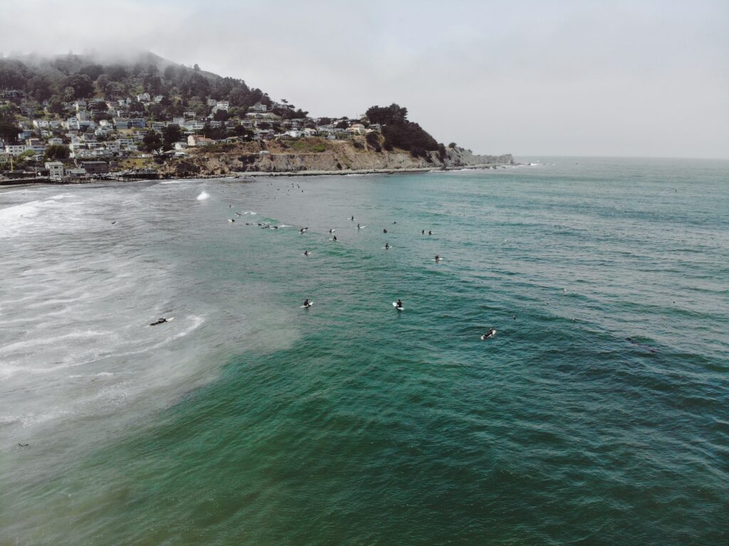 Surfers sit in the green-blue water at Pacifica surf State Beach, California, waiting for a wave, with cliffs and houses perched above the shoreline in the background