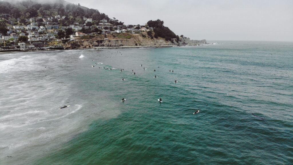 Surfers sit in the green-blue water at Pacifica surf State Beach, California, waiting for a wave, with cliffs and houses perched above the shoreline in the background