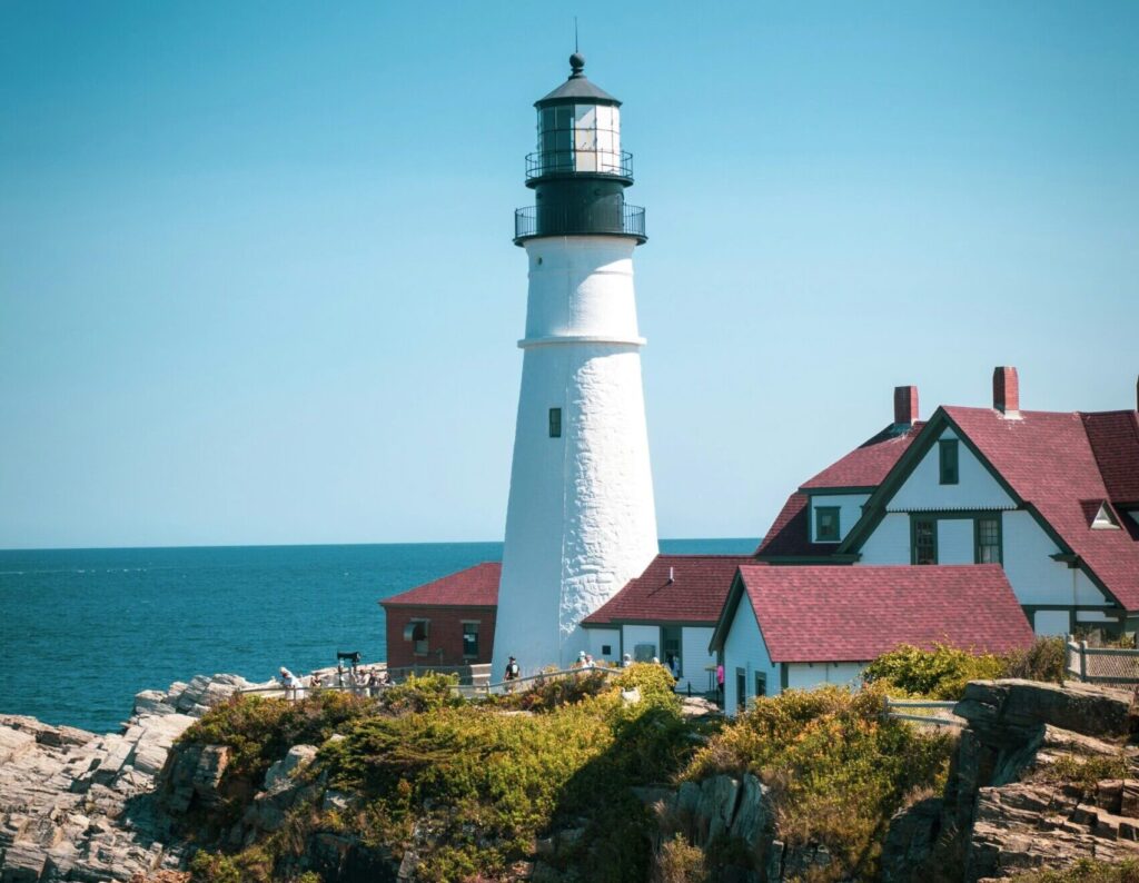 Portland Head Lighthouse standing tall against a backdrop of clear blue skies, with nearby houses featuring red roofs nestled along the coastline.