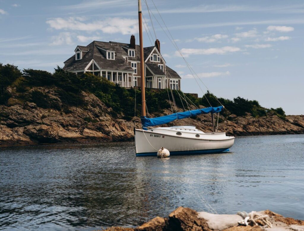 A small yacht floats peacefully in the harbor at Newport, Rhode Island, with a large brown house in the background and clear, cloudy skies above