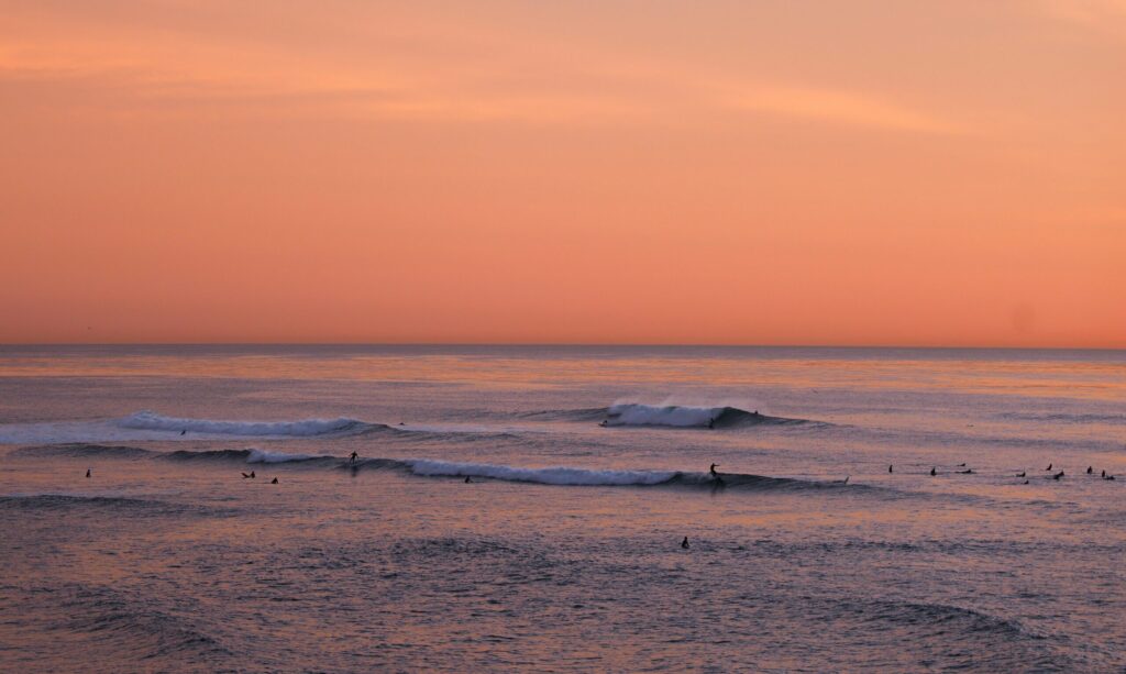 Surfers in the distance catching waves at Sunset Cliffs surf, CA, under vibrant orange skies as the sun sets over the ocean