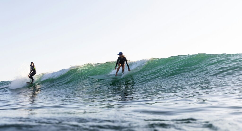 A surfer dropping in on another rider on a left-hand wave under clear skies with vibrant green-blue water in El Palmar Surf, Spain