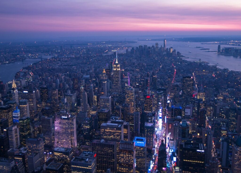 Drone shot of Manhattan, New York, with city lights glowing as the sun sets, casting a dim, evening light over the skyline