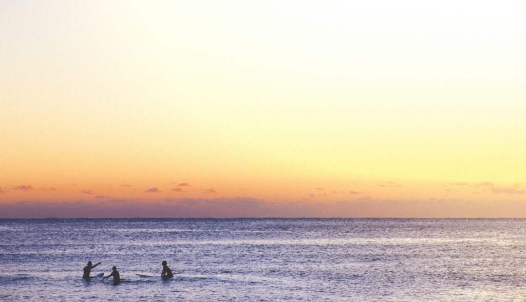 Three surfers chatting in calm ocean water as the sun sets, casting soft yellow-white tones across the sky in El Palmar surf, Spain