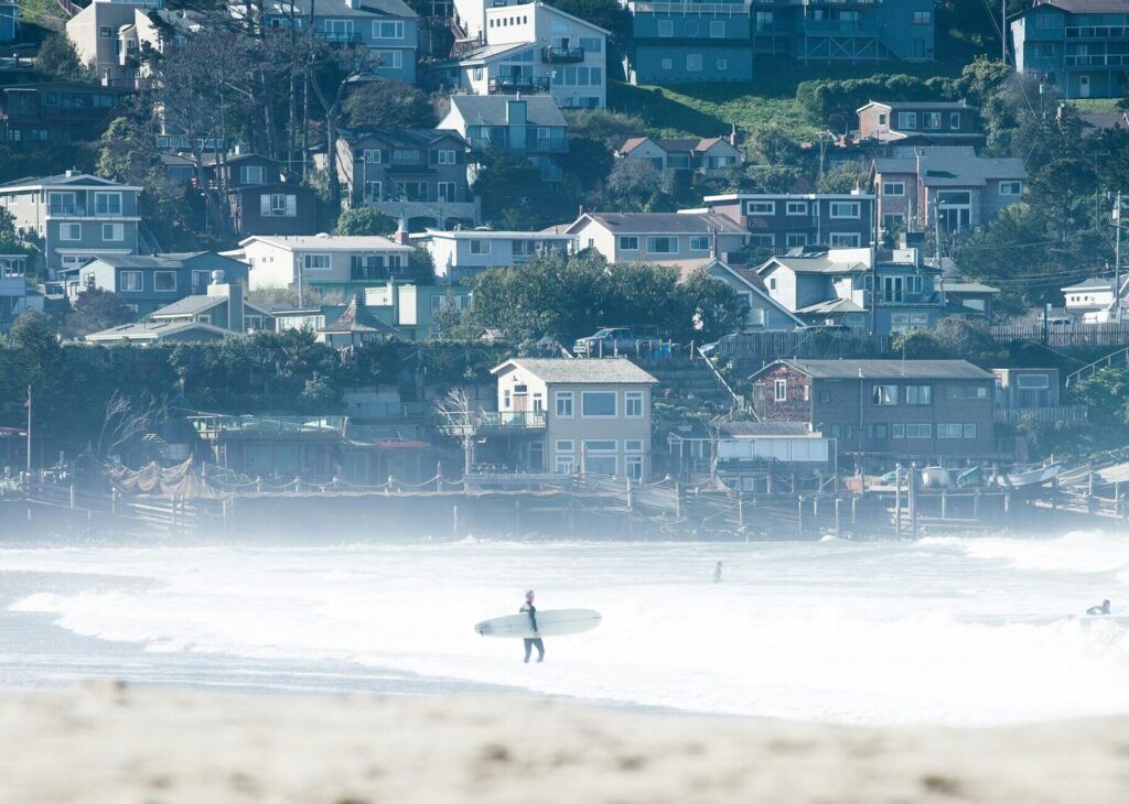 A surfer is seen from a distance entering the water at Pacifica surf State Beach, California, with houses lining the backdrop against the coastal landscape