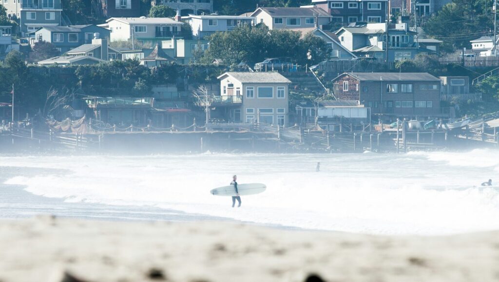 A surfer is seen from a distance entering the water at Pacifica surf State Beach, California, with houses lining the backdrop against the coastal landscape