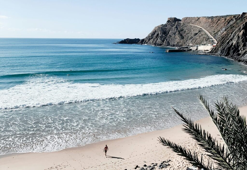 A man walks out of the blue ocean at Arrifana, Algarve, with the sun shining and gentle waves lapping at the shore