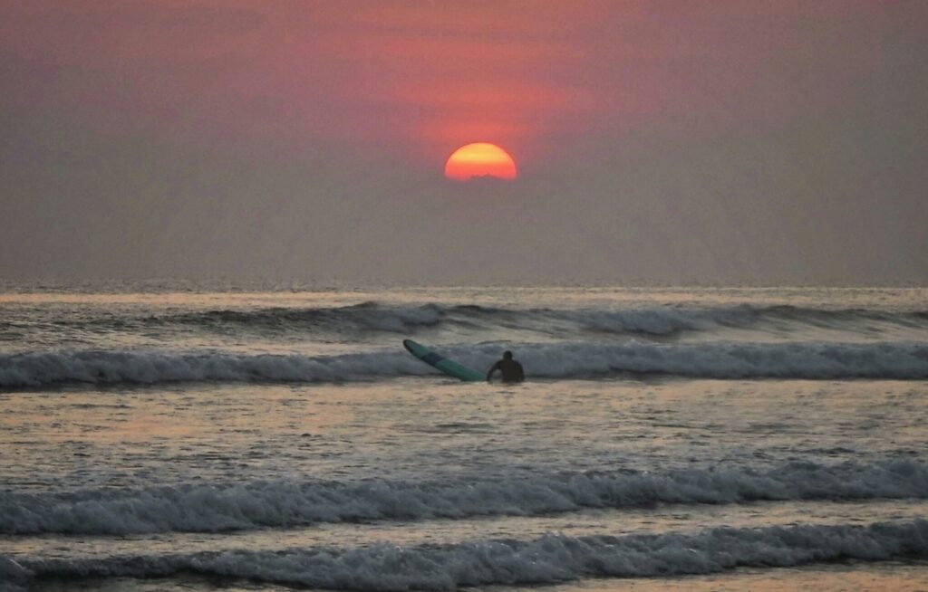 Seminyak surf sunset in Indonesia, showing a surfer sitting on a board with red skies over the ocean