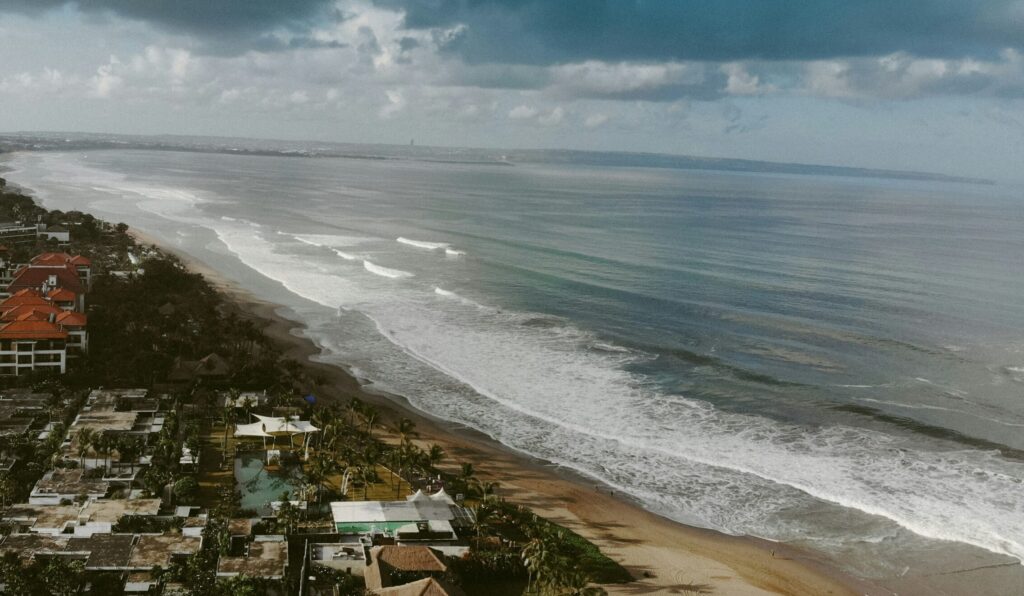 Drone view of Seminyak surf Beach, Indonesia, with waves and beach on the right, houses and greenery on the left