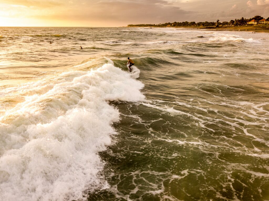 Surfer rides a long left-hand wave at Canggu Surf Beach, Indonesia, under yellow skies with a shimmering water reflection