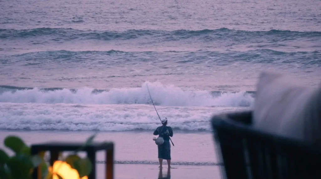 Man fishing at Seminyak Beach, Indonesia, with large waves in the background and light red cloudy skies