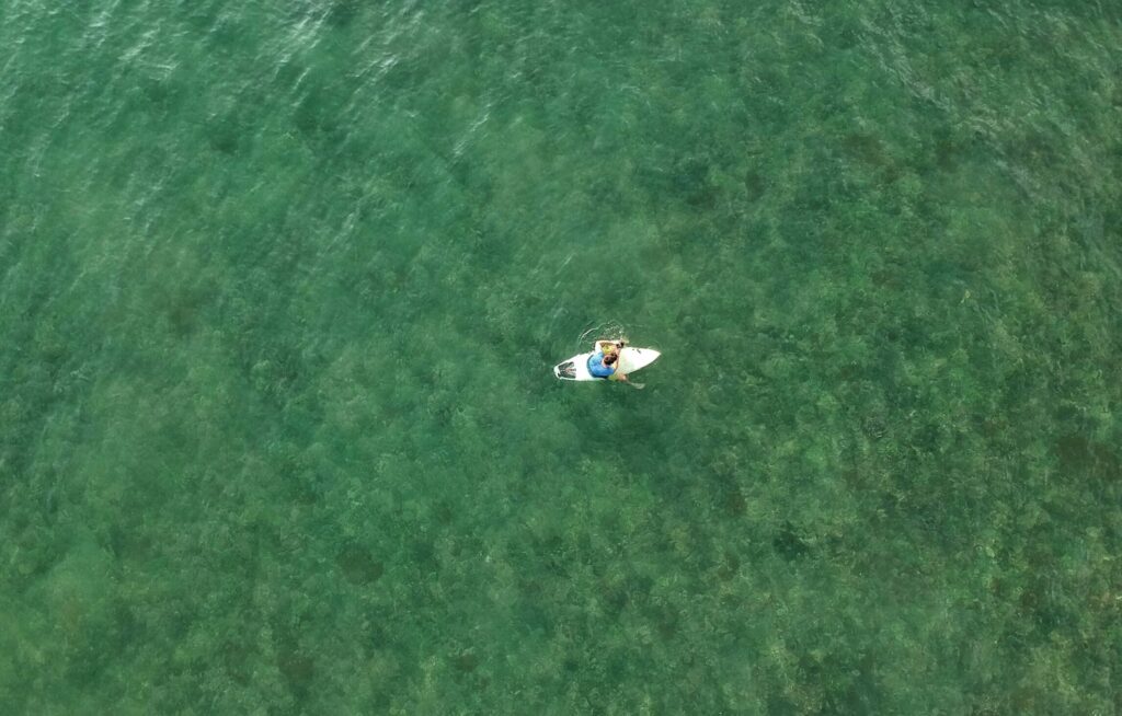 Drone view of a surfer sitting on a board in clear blue-green water, waiting for a wave at Cimaja Surf Beach