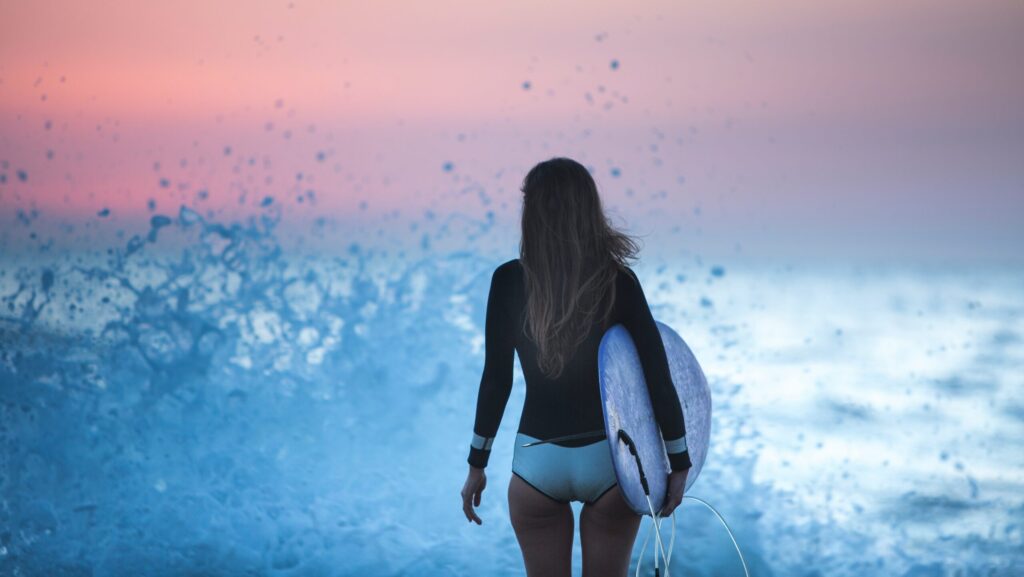 A surfer girl holding a surfboard in Troncones surf, looking out into the ocean with red skies in the background