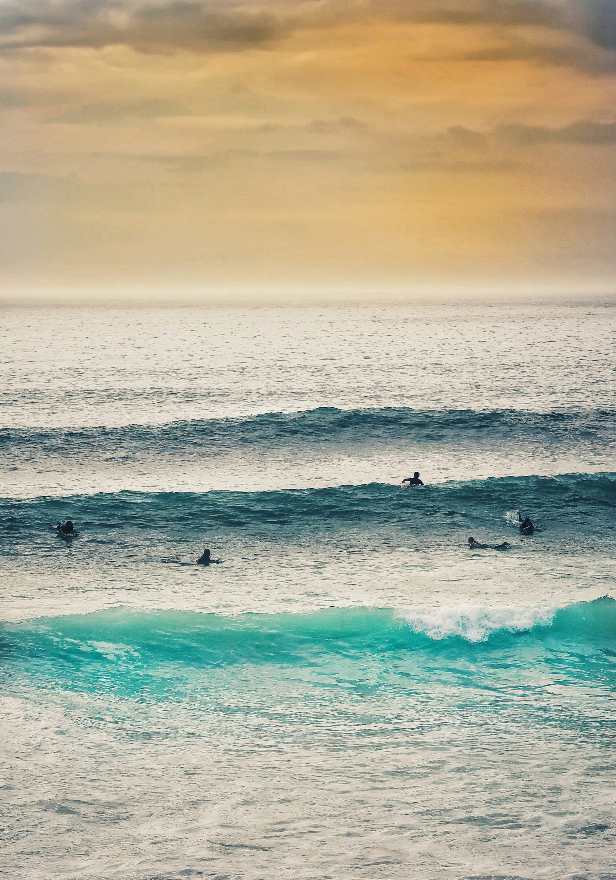 Surfers at Fistral Beach, Newquay surf, with a two-wave set, one paddling into a wave under low sun and yellow skies