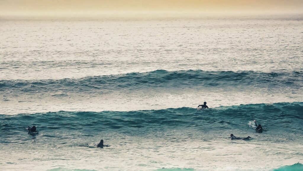Surfers at Fistral Beach, Newquay surf, with a two-wave set, one paddling into a wave under low sun and yellow skies