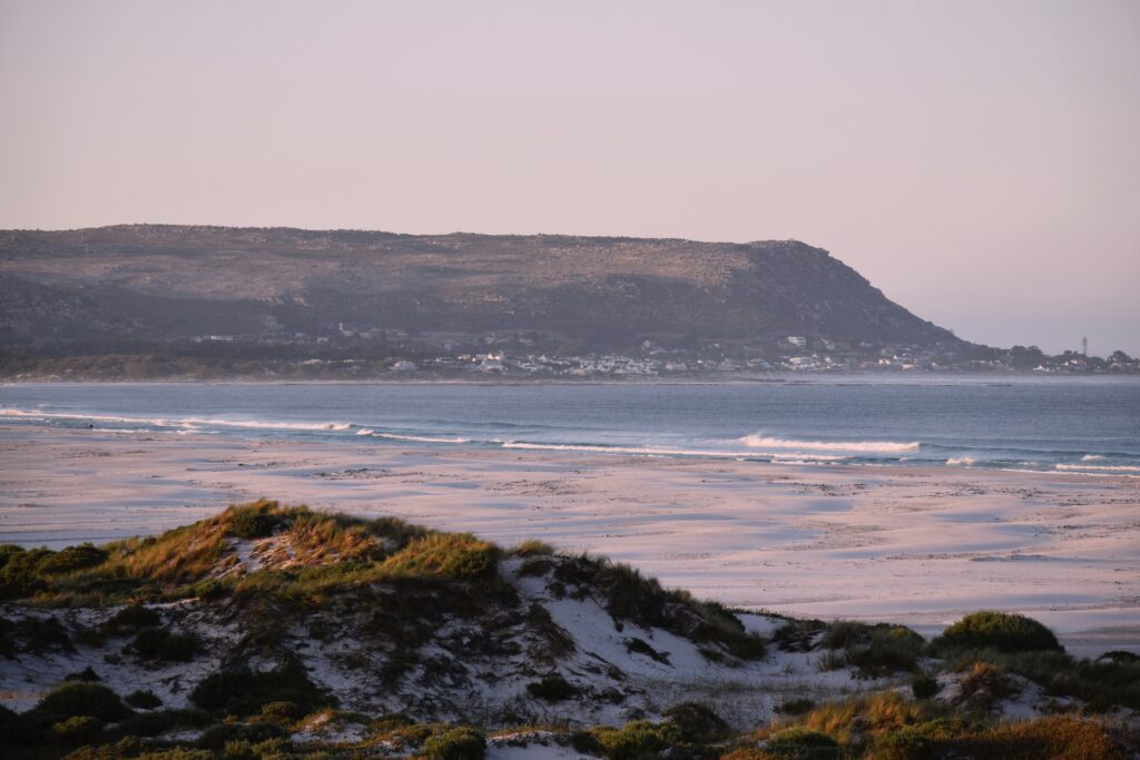 Sandy foreground with waves in the distance, mountains, greenery, and clear blue skies at Noordhoek, in Kommetjie Surf.