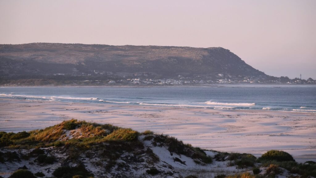 Sandy foreground with waves in the distance, mountains, greenery, and clear blue skies at Noordhoek, in Kommetjie Surf.