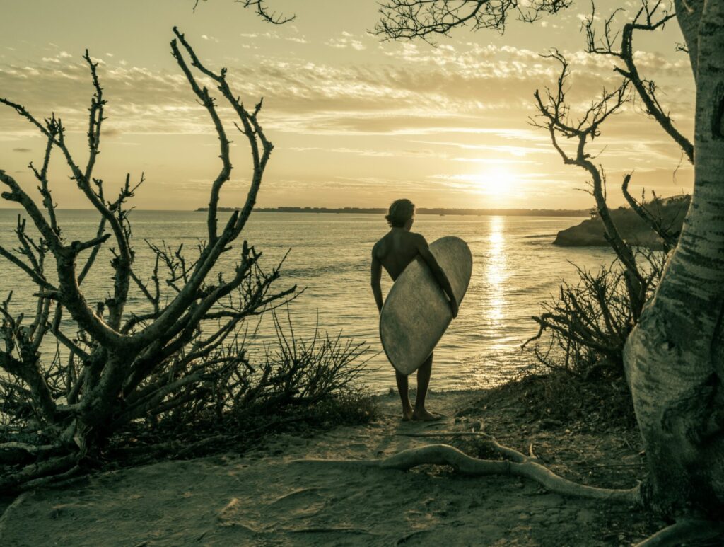 A surfer holding a longboard, looking out at Sayulita surf Beach, Mexico, in black and white, with the beach in the background