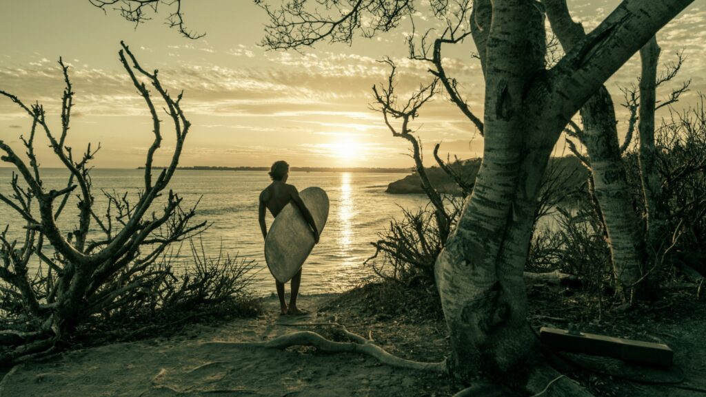 A surfer holding a longboard, looking out at Sayulita surf Beach, Mexico, in black and white, with the beach in the background