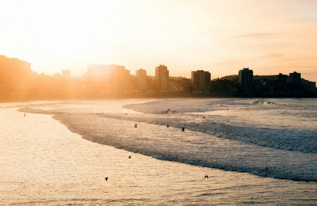 A side view of the beach and surf in Gijon Surf, Asturias, with the city in the background as the sun sets, creating a bright yellow tinge in the sky