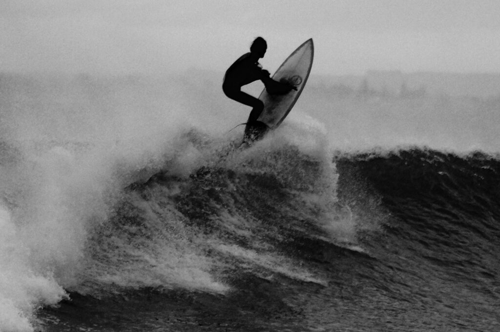 A surfer holding the board while executing an aerial move on a left-hand wave in black and white in Mundaka Surf, Spain