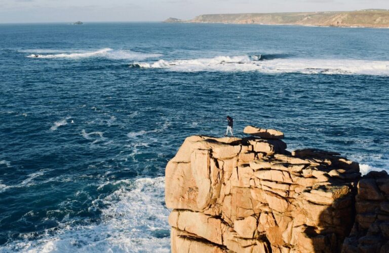 A person standing on a cliff with a wave breaking on a reef in the background under clear blue skies