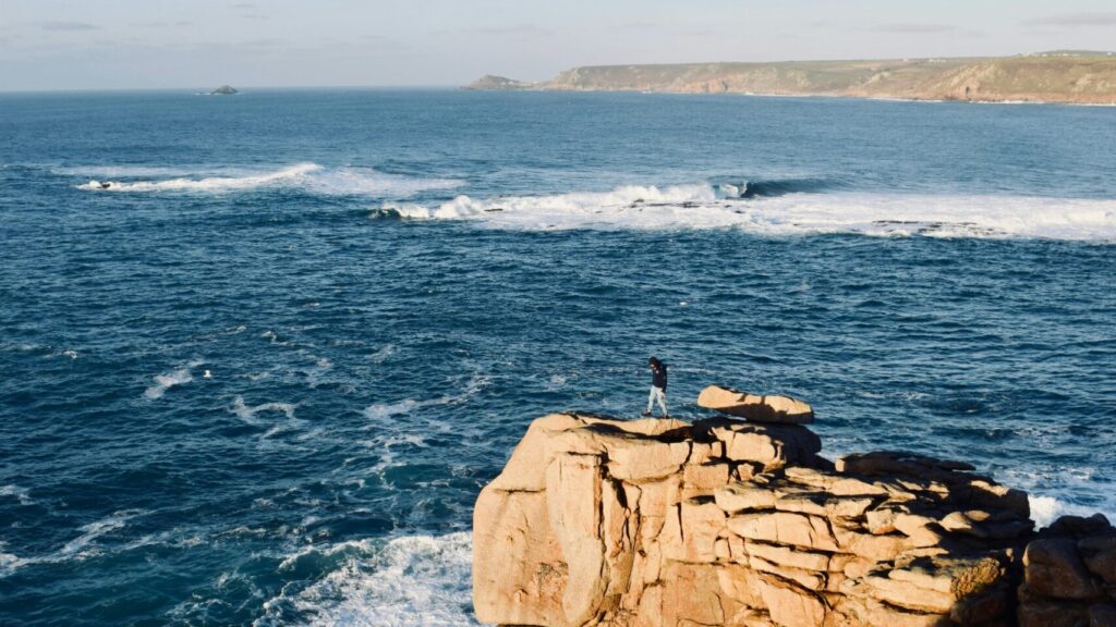 A person standing on a cliff with a wave breaking on a reef in the background under clear blue skies