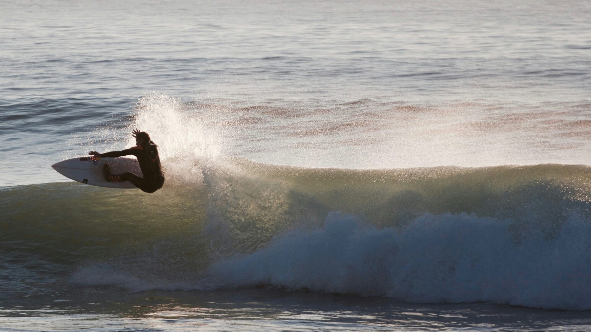A surfer performing a powerful top turn on a right-hand wave, with sunlight reflecting on the water in Bantham Surf, United Kingdom