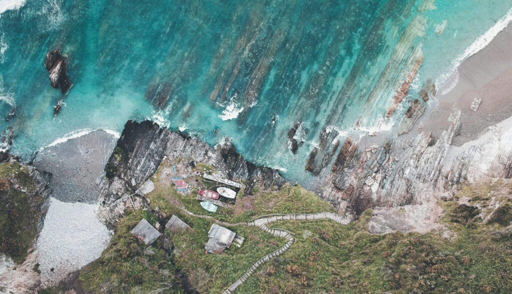 A drone's eye view of a cliff with stairs leading down to crisp, clear blue water in Gijon, Asturias, featuring rocky formations in the water in Gijon Sur