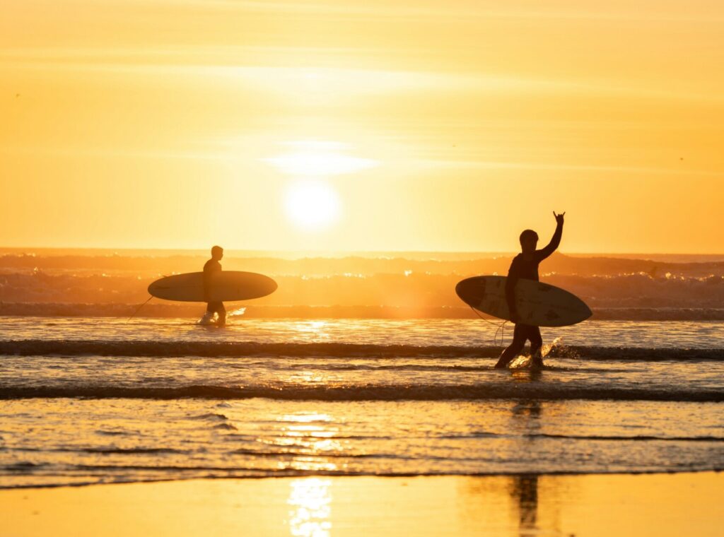 Two surfers walk out of the water at Cimaja Beach, bathed in warm yellow sunlight.