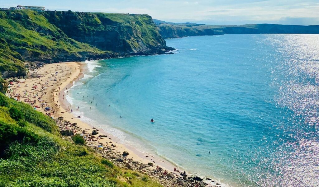 Beach at Suances, Spain, with people on the sand and swimming, clear blue flat water, greenery, and cliffs in the background